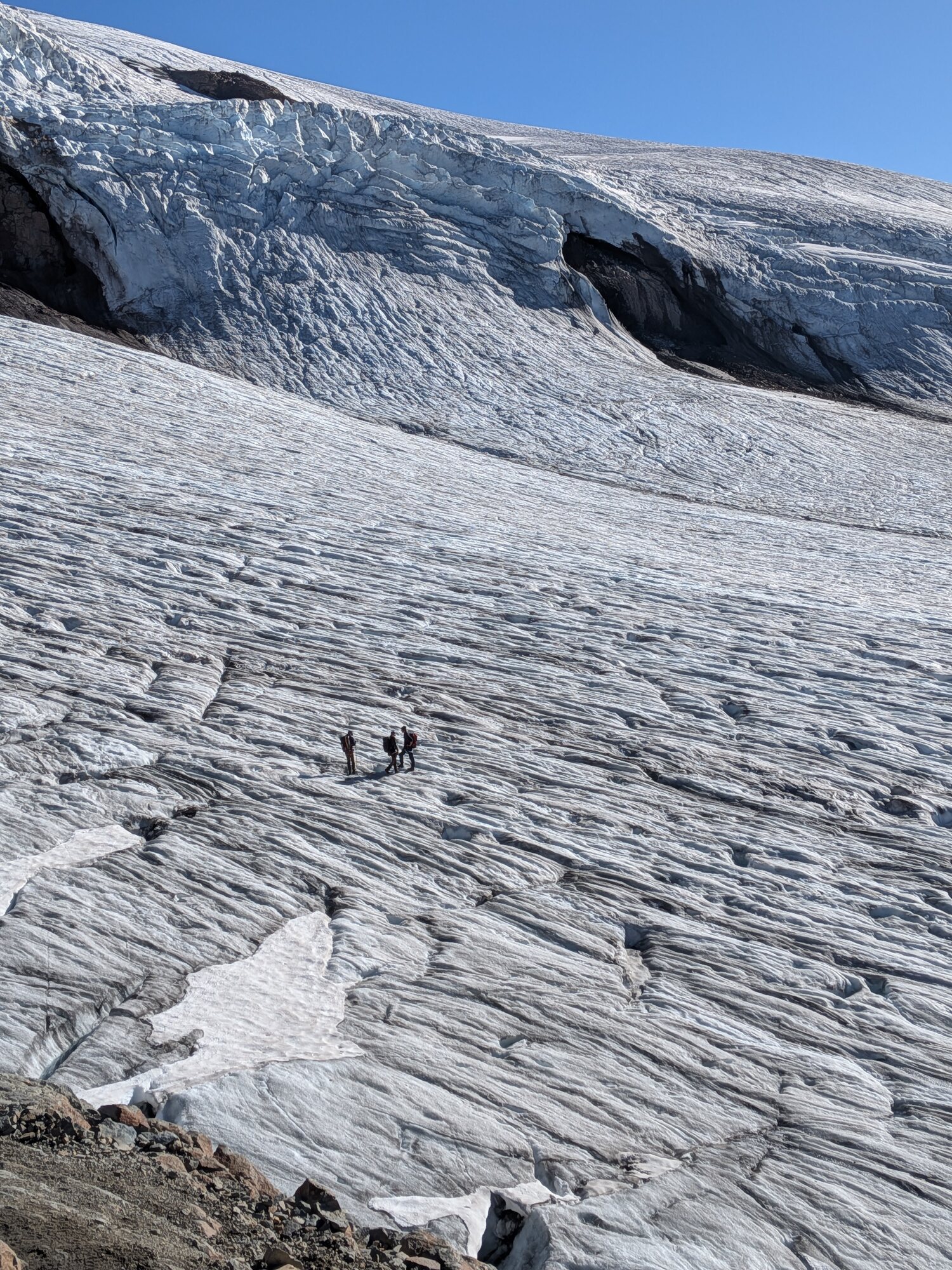 Three field researchers crossing a fractured glacier beneath a heavily crevassed ice slope